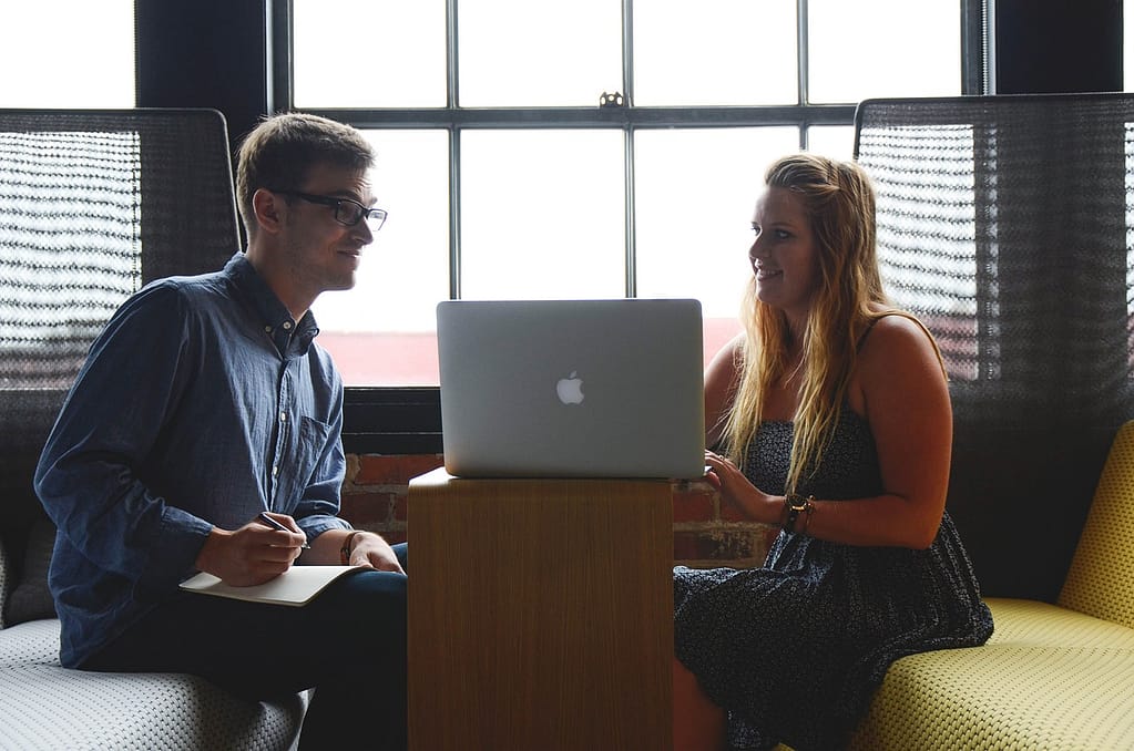 Pareja revisando información en un portátil Mac frente a una ventana, representando colaboración y comunicación.